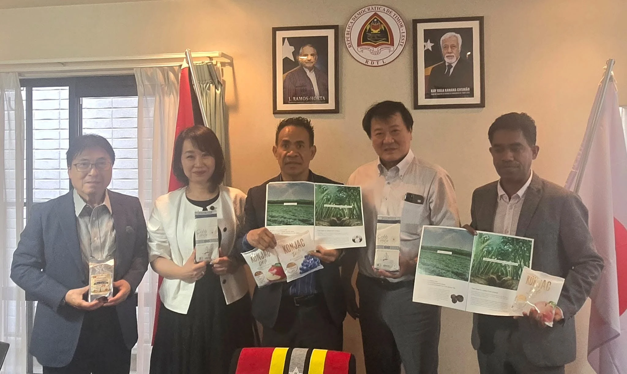 Group photo at the Embassy of Timor-Leste in Japan holding maek-based konjac products and Café Timor packaging, beneath portraits of President Ramos-Horta and Kay Rala Xanana Gusmão